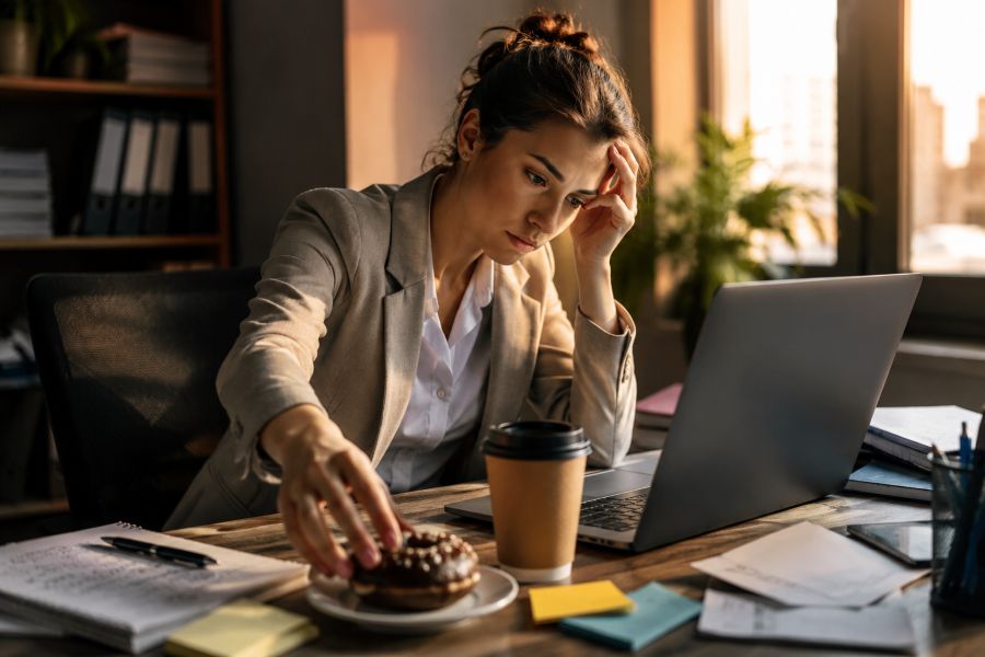 Tired professional reaching for a sugary snack and coffee while working late, representing the connection between stress hormones, cravings, and unstable energy.